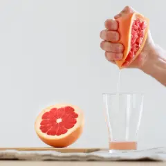 Ręka wyciska soczystego grejpfruta, a jego świeży sok kapie do szklanki. Obok leży połówka owocu.