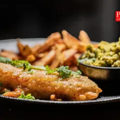 Chrupiące fish and chips z zielonym groszkiem i świeżą miętą na czarnym talerzu.
