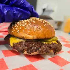 Rękawiczka w niebieskiej rękawicy dociska idealnie wysmażonego smash burgera z serem i ogórkiem. Oto jak wygląda smash burger co to!