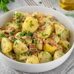 Pyszny kartoffelsalat przepis: ziemniaki z boczkiem, cebulką i ziołami w białej misce, obok cytryna i oliwa.