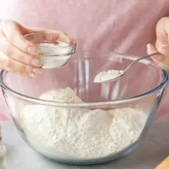 Kobieta dodaje sodę oczyszczoną do mąki w szklanej misce. Szukasz co zamiast proszku do pieczenia? Soda jest świetnym zamiennikiem!