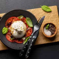 Pyszna burrata z pomidorami, polana oliwą, udekorowana bazylią. Obok miseczka z solą i pieprzem.