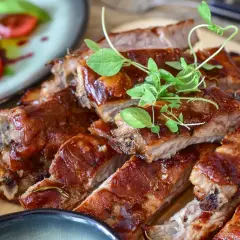 Soczyste żeberka w sosie BBQ, doprawione ziołami, wyglądają apetycznie. Obok talerz z pomidorami i bazylią.