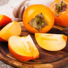 Pomarańczowe, soczyste kaki pokrojone w ćwiartki i całe owoce leżą na drewnianej desce.