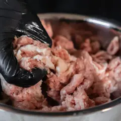 Ręka w czarnej rękawiczce miesza surowe mielone mięso w metalowej misce, przygotowując składniki na obiad.