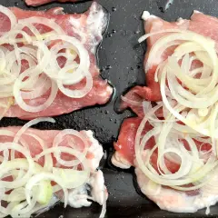 Karkówka w plastrach z cebulą na czarnej blasze. Idealny pomysł na karkówkę, która zaraz trafi do piekarnika.