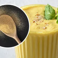 Puszysty mango lassi w szklance, ozdobiony miętą i orzechami. Obok łyżka z proszkiem, być może przyprawą do mango lassi.