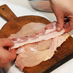 Ręce układają plastry boczku na piersi z kurczaka, przygotowując pyszne danie: pierś z kurczaka zawijana w boczku z serem.