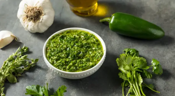 Miseczka pełna aromatycznego sosu chimichurri, obok świeża kolendra, natka pietruszki, czosnek, papryczka jalapeno i oliwa.