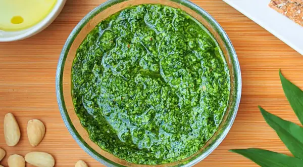 Aromatic pesto z czosnku niedźwiedziego w szklanej miseczce, otoczone migdałami i świeżymi liśćmi.