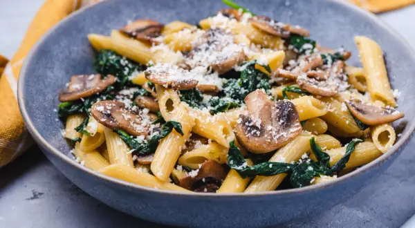 Penne z pieczarkami i szpinakiem, posypane parmezanem, w szarej misce.