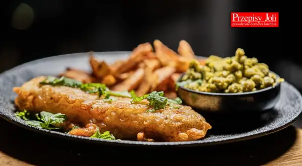 Chrupiące fish and chips z zielonym groszkiem i świeżą miętą na czarnym talerzu.