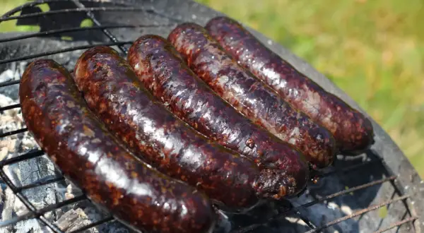 Pięć ciemnych, błyszczących kiełbasek kaszanka na grillu, dymiących na ruszcie nad żarem.