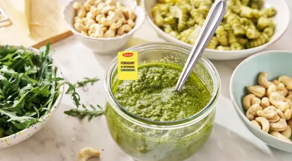 Pesto z rukoli w słoiku, obok miseczki z orzechami nerkowca i rukolą. W tle makaron z zielonym sosem.