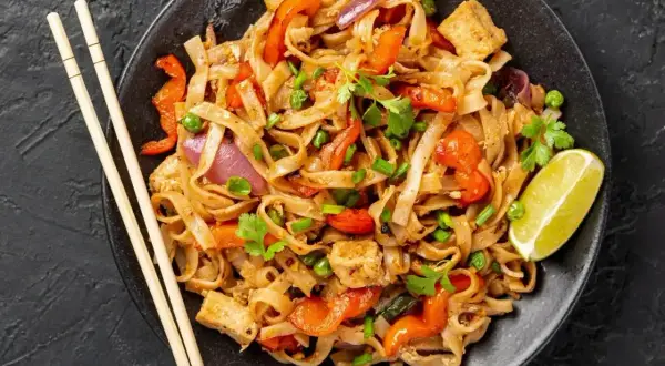 Talerz pełen makaronu ryżowego z warzywami i tofu, polany aromatycznym sosem pad thai. Obok pałeczki i ćwiartka limonki.