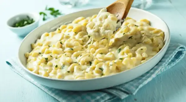 Pyszny makaron z mozzarellą i ziołami, podany w białej misie. Drewniana łyżka nabiera porcję kremowego dania.