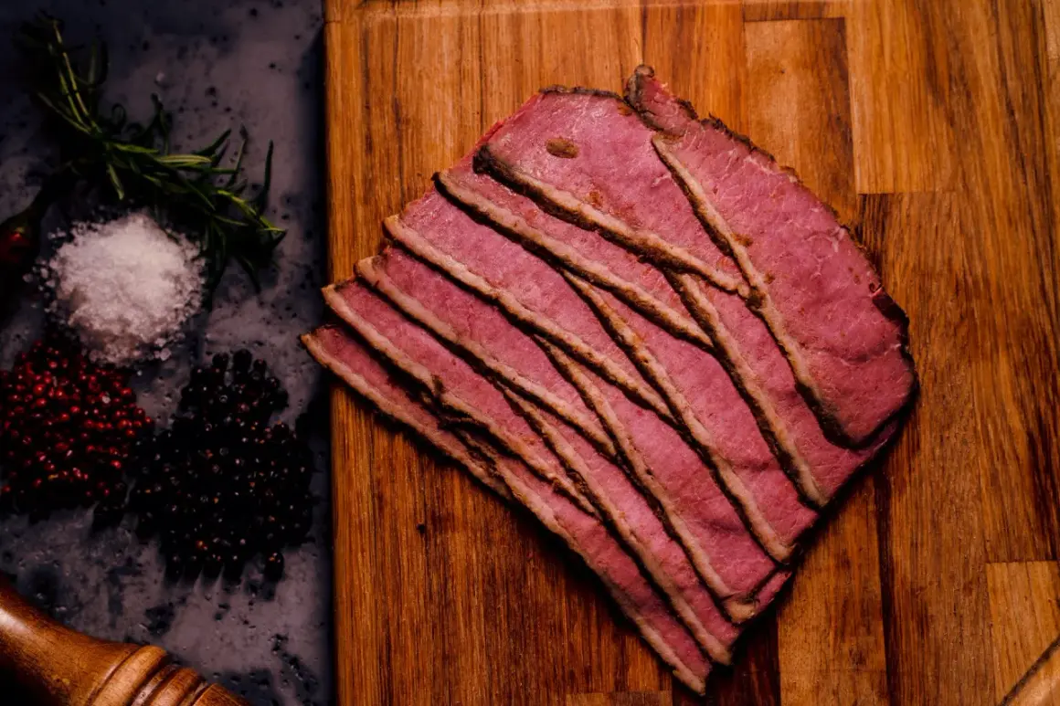 Cienkie plastry różowego pastrami na desce, obok przyprawy. Zastanawiasz się, pastrami co to? To aromatyczna, peklowana wołowina.