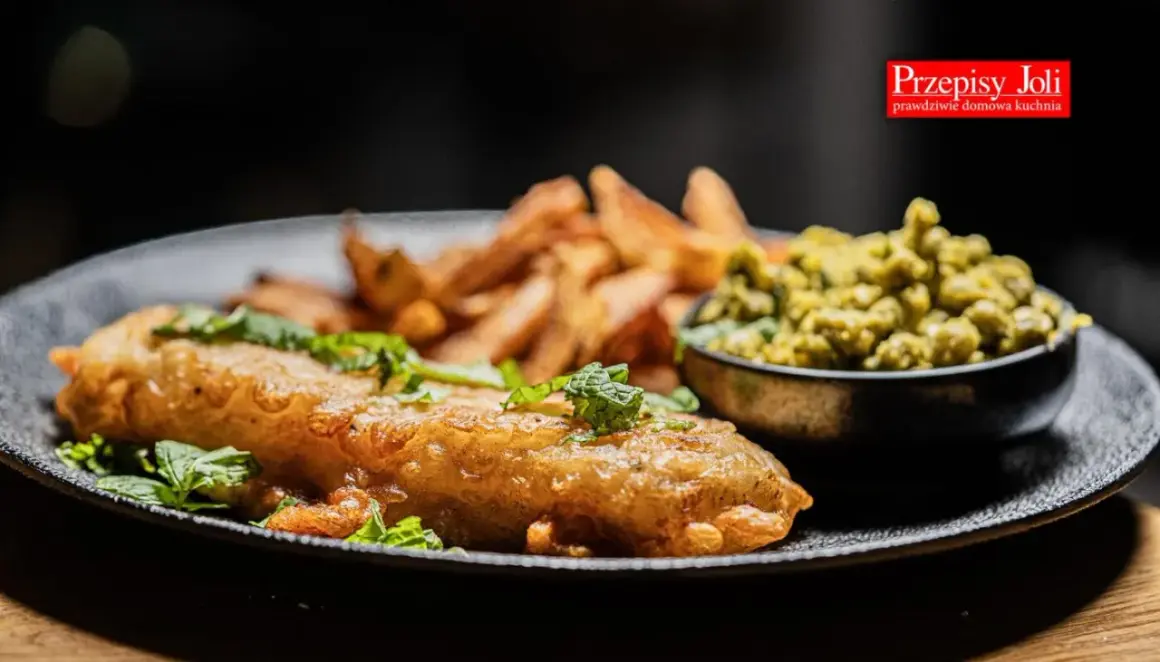 Chrupiące fish and chips z zielonym groszkiem i świeżą miętą na czarnym talerzu.