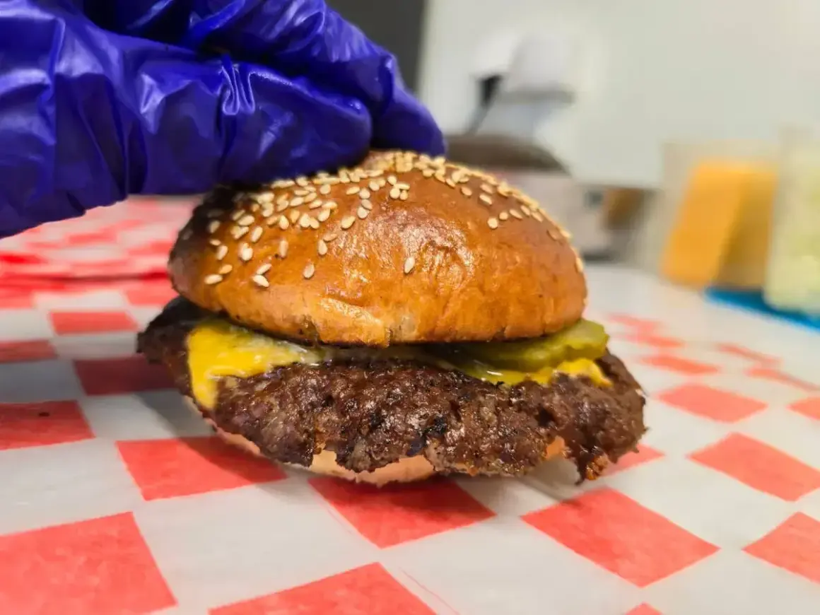 Rękawiczka w niebieskiej rękawicy dociska idealnie wysmażonego smash burgera z serem i ogórkiem. Oto jak wygląda smash burger co to!