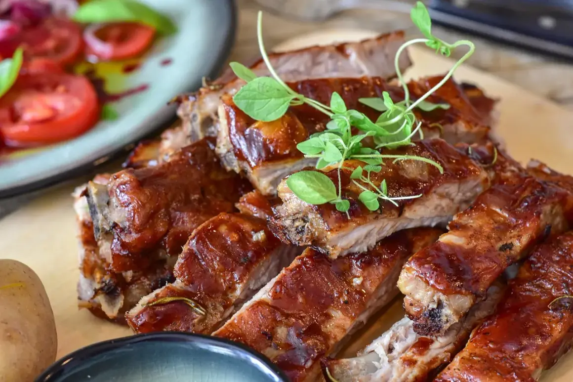 Soczyste żeberka w sosie BBQ, doprawione ziołami, wyglądają apetycznie. Obok talerz z pomidorami i bazylią.