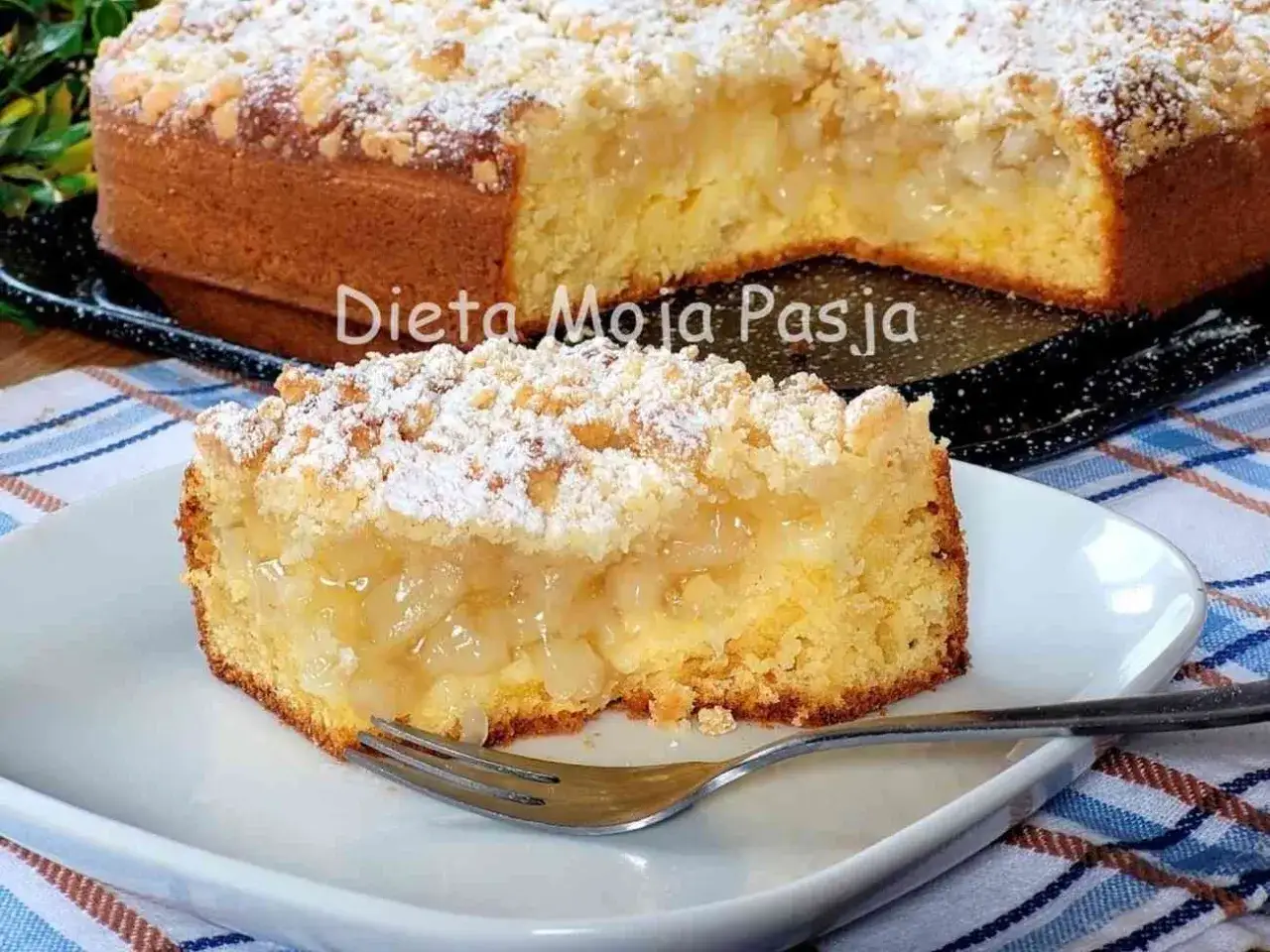 Wilgotne ciasto z jabłkami na oleju, posypane kruszonką i cukrem pudrem. Pyszny kawałek czeka na widelcu.