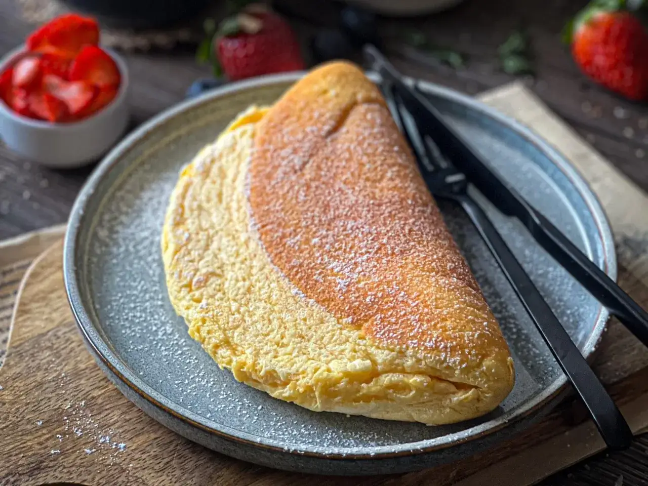 Puszysty omlet z mąką, posypany cukrem pudrem, czeka na śniadanie. Obok miseczka z truskawkami i sztućce.