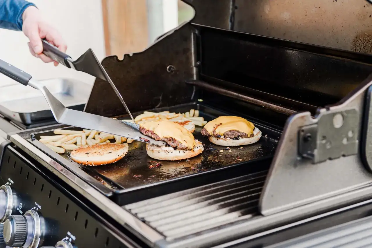 Przygotowanie smash burgerów na grillu. Mięso jest rozgniatane, a na nim topiony ser. Obok smażą się frytki.